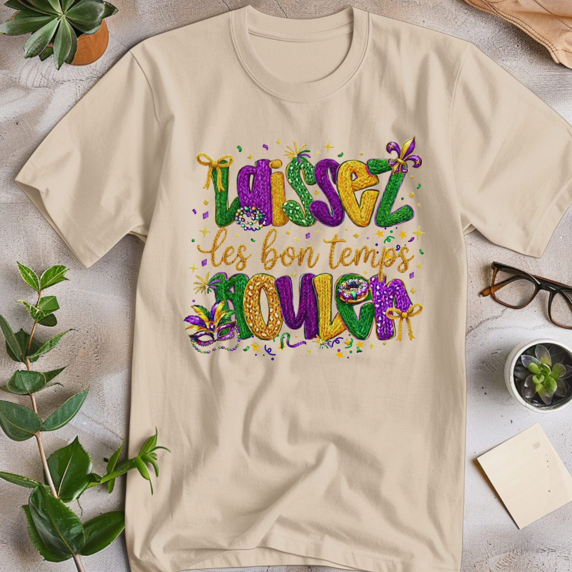 Beige t-shirt with colorful text design on a light surface with plants and glasses.