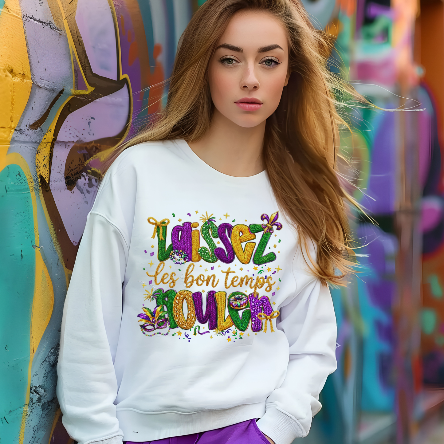 Woman wearing a white sweatshirt with colorful text in front of a graffiti wall.