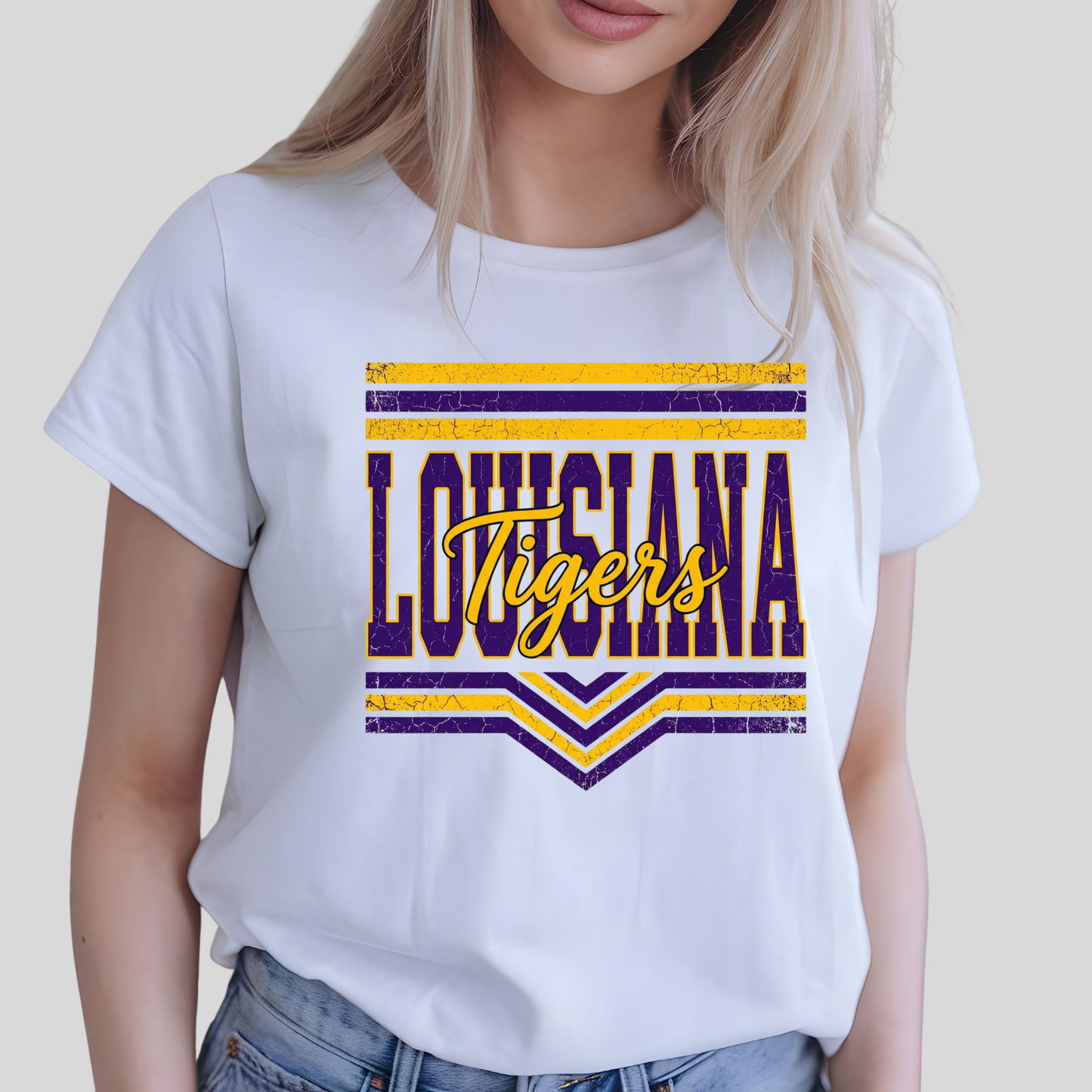A woman wearing a white short-sleeve graphic T-shirt with a distressed purple and yellow 'LOUISIANA Tigers' design on the front, paired with blue jeans against a light gray background.