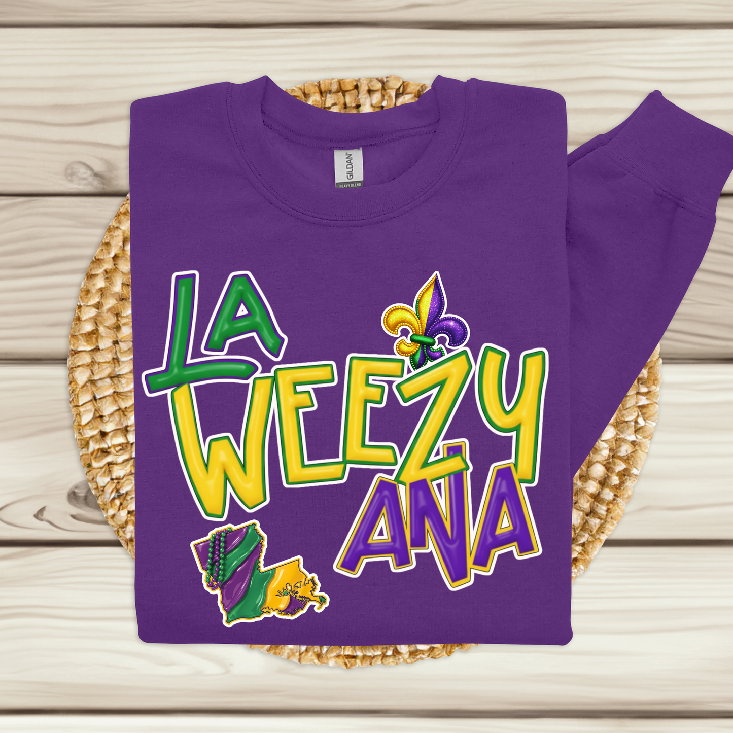 Purple sweatshirt with 'La Weezy Ana' text and Mardi Gras-themed design on a woven mat.