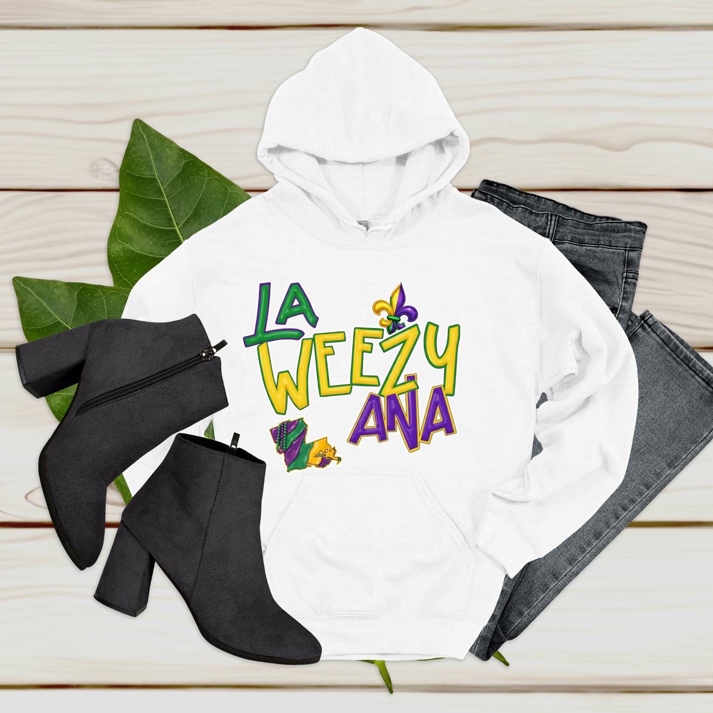 White hoodie with 'La Weezy Ana' text, black boots, and jeans on a wooden surface
