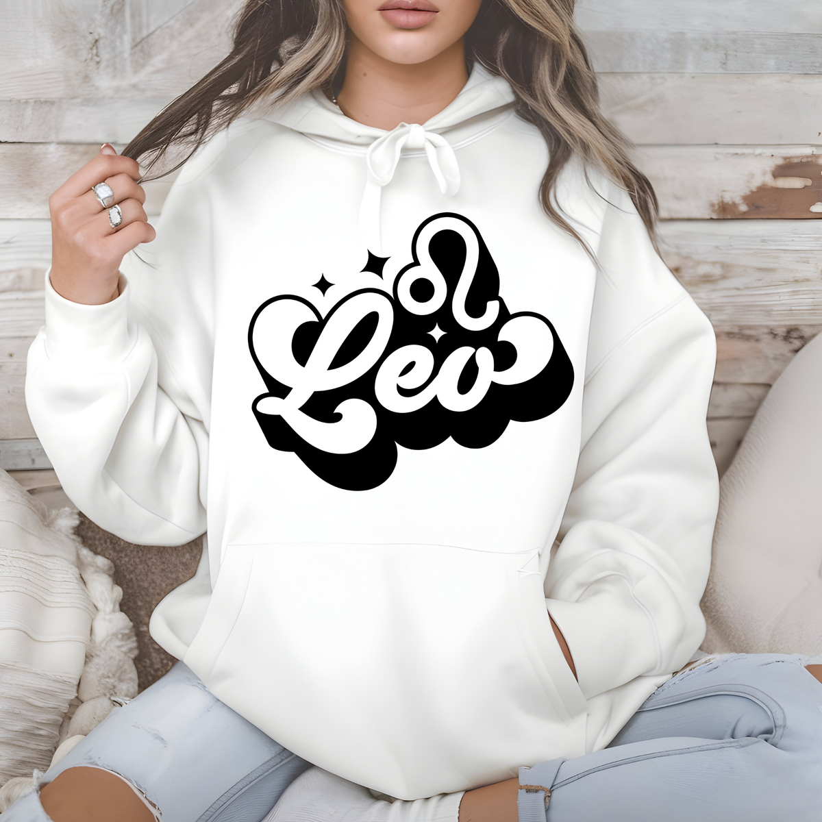 A woman wearing a white pullover hoodie with a black 'm Virgo' graphic on the chest, sitting against a rustic wood and neutral background.