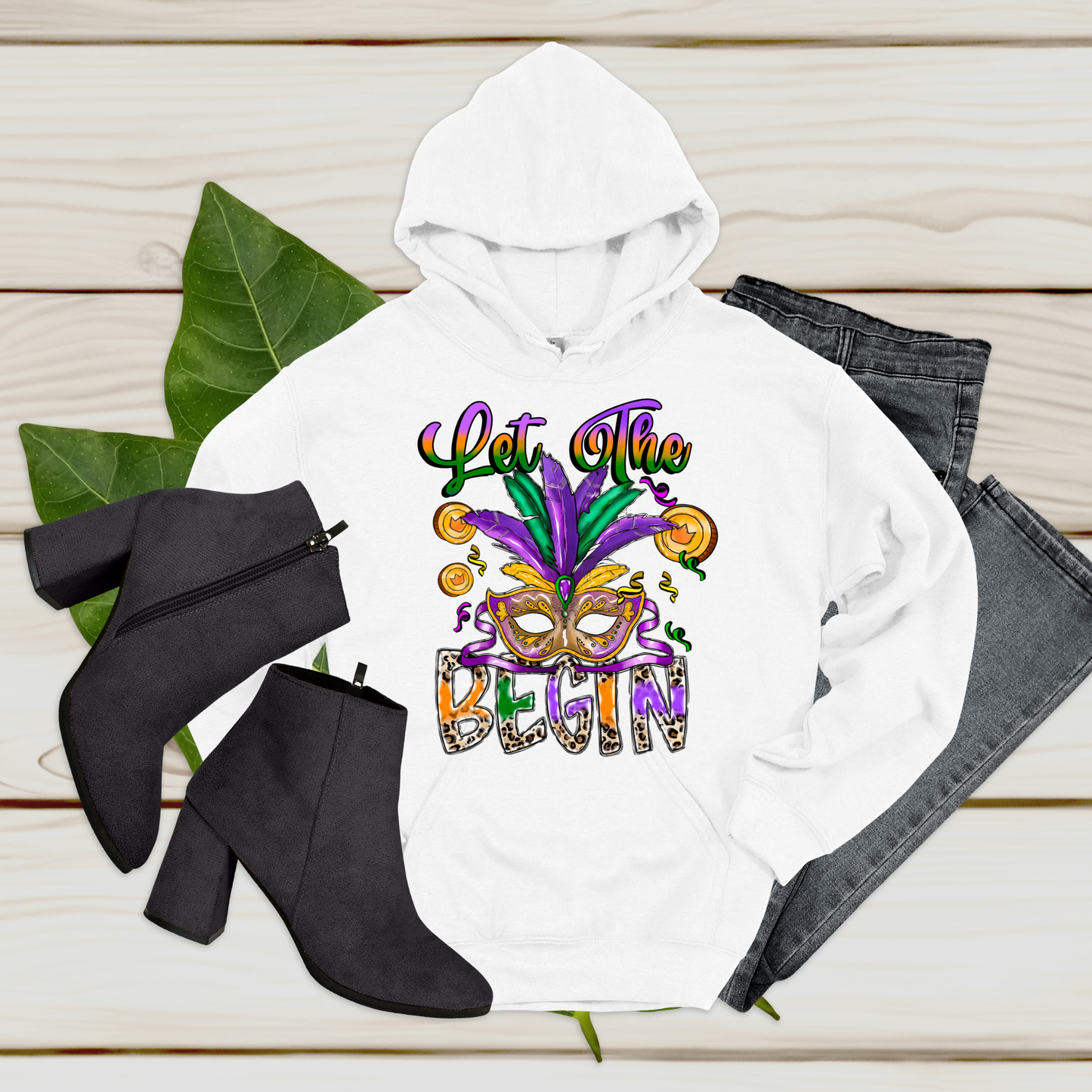 White hoodie with Mardi Gras-themed design, black boots, and jeans on a wooden surface.