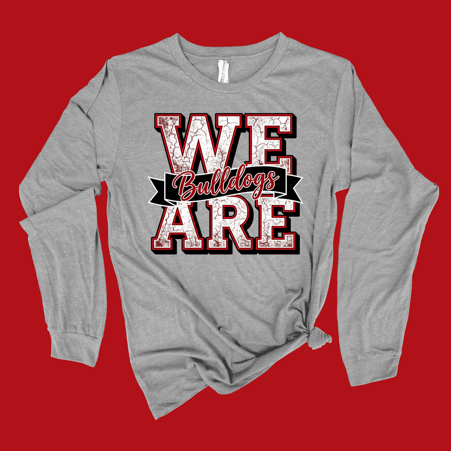A heather gray long-sleeve t-shirt with a distressed graphic reading 'WE Bulldogs ARE' in red, white, and black, styled with a knot at the bottom hem, set against a solid red background.