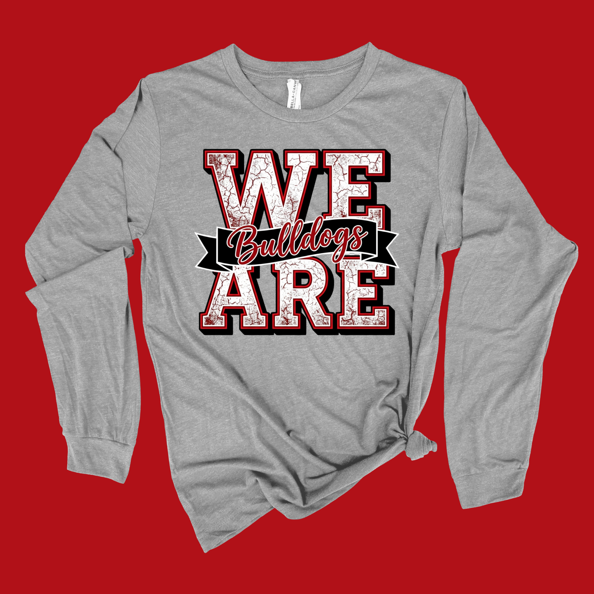 A heather gray long-sleeve t-shirt with a distressed graphic reading 'WE Bulldogs ARE' in red, white, and black, styled with a knot at the bottom hem, set against a solid red background.