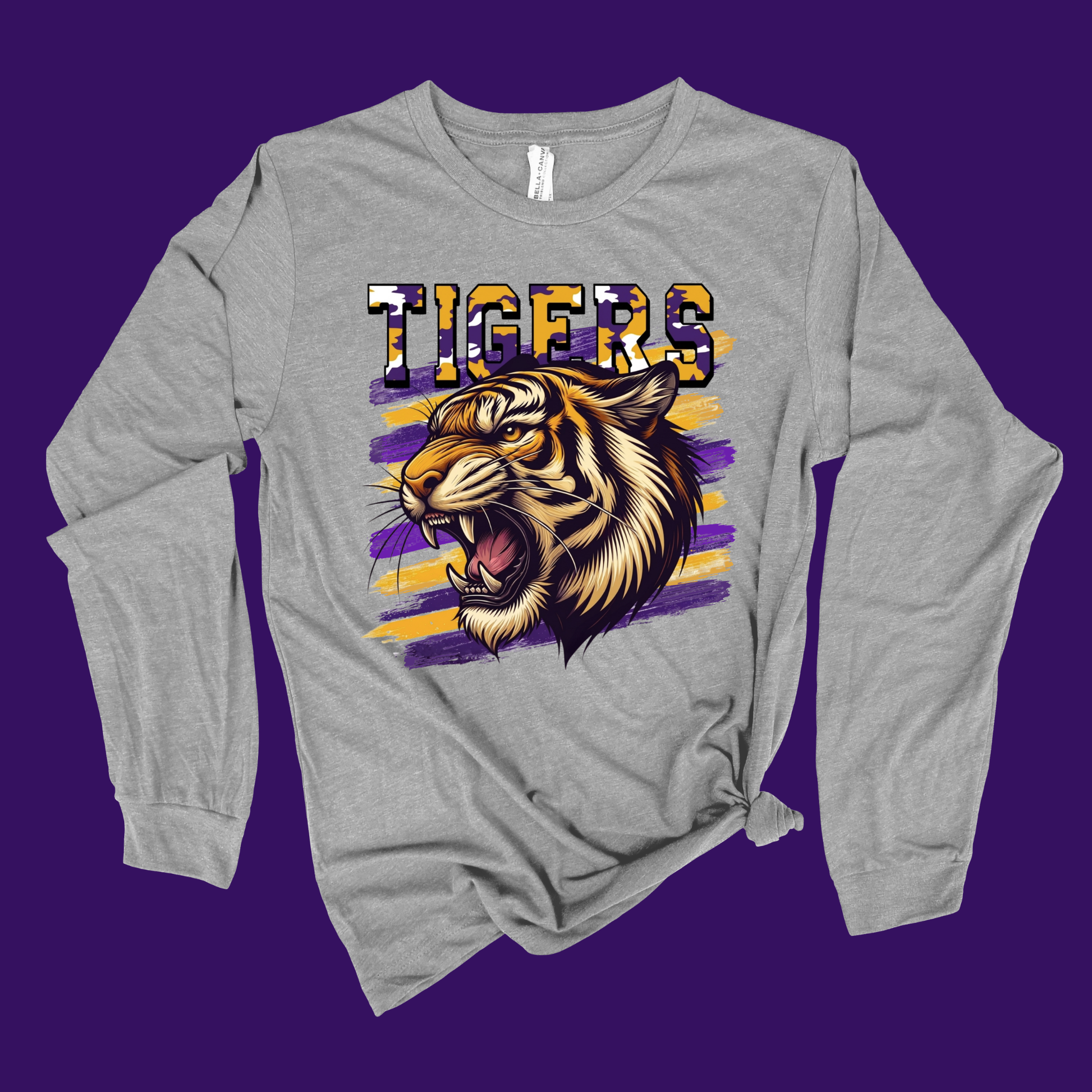 A heather gray long-sleeve t-shirt with a graphic of a roaring tiger and the word 'TIGERS' in a patriotic flag pattern, set against a solid purple background.