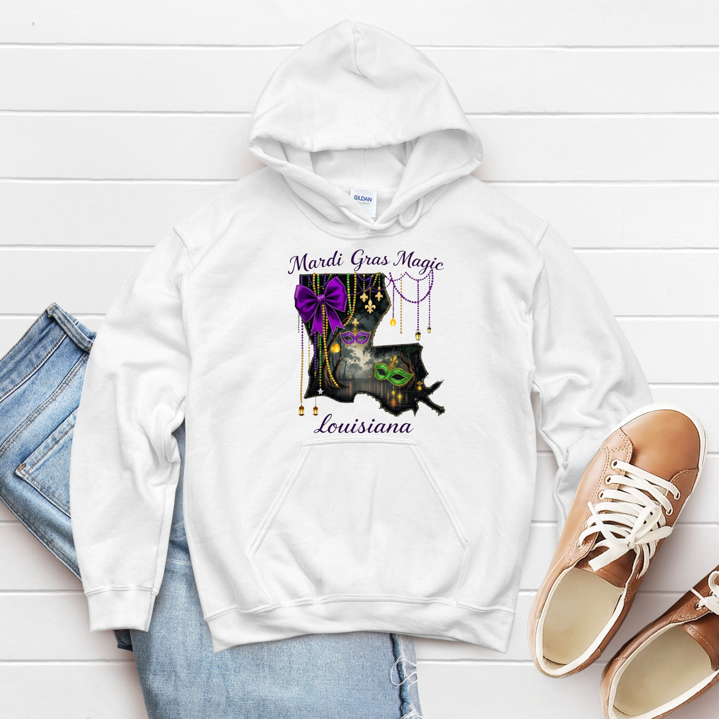 White hoodie with Mardi Gras-themed design on a white wooden surface with jeans and shoes.