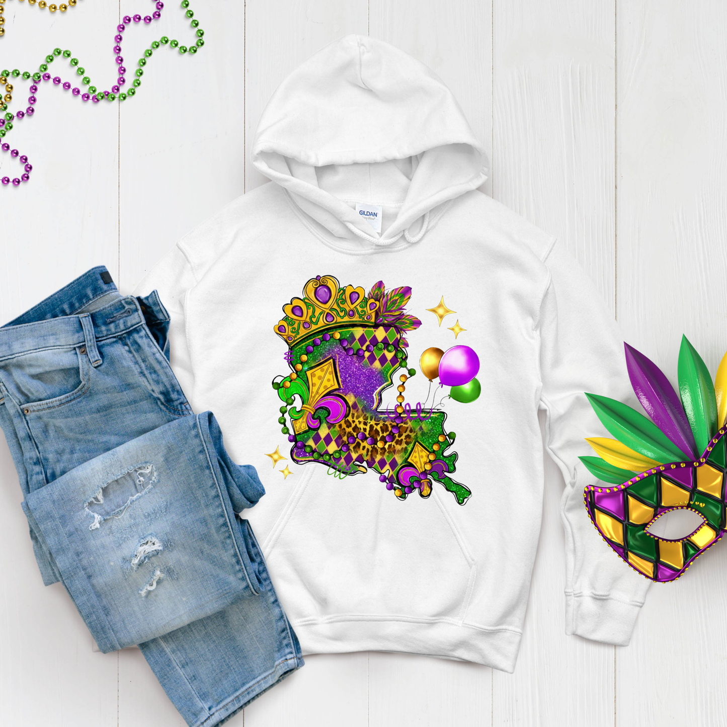 White hoodie with Mardi Gras-themed design, jeans, and mask on a light wooden surface.