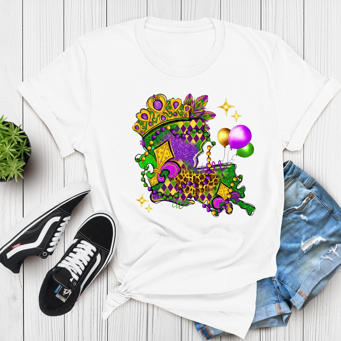 White t-shirt with colorful Mardi Gras design, black sneakers, and denim shorts on a wooden surface.