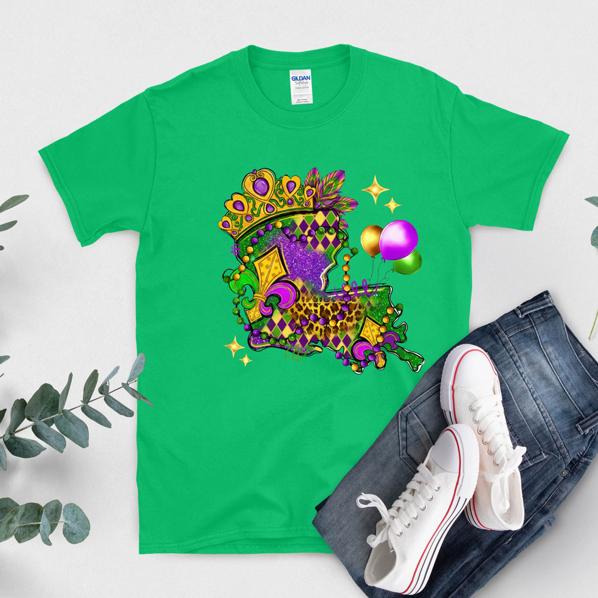 Green t-shirt with Mardi Gras-themed design, white sneakers, and jeans on a light gray background.