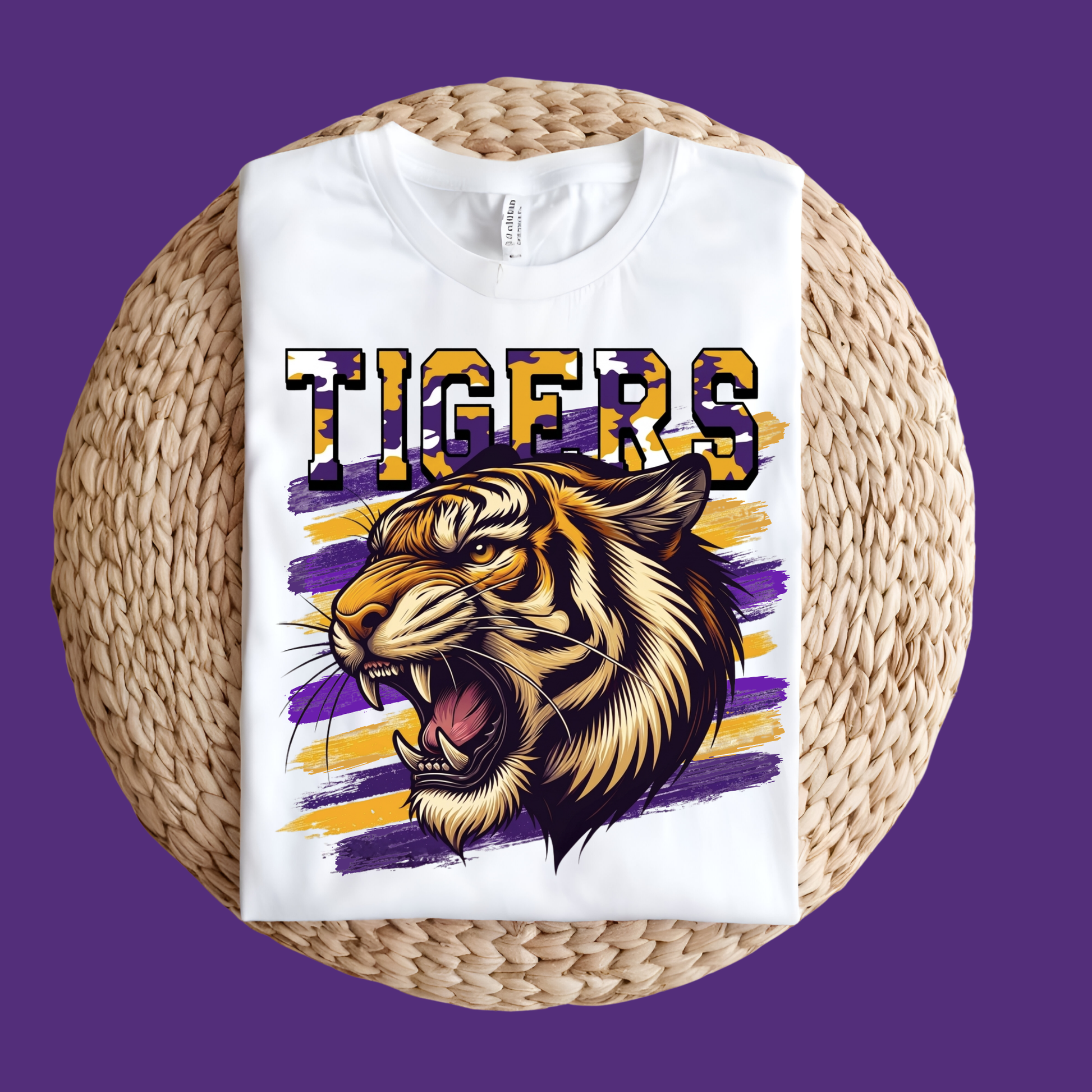 A white short-sleeve T-shirt laid flat on a round woven straw mat, featuring a large graphic of a roaring tiger head and the word "TIGERS" in purple, gold, and black. The shirt is photographed against a solid purple background.