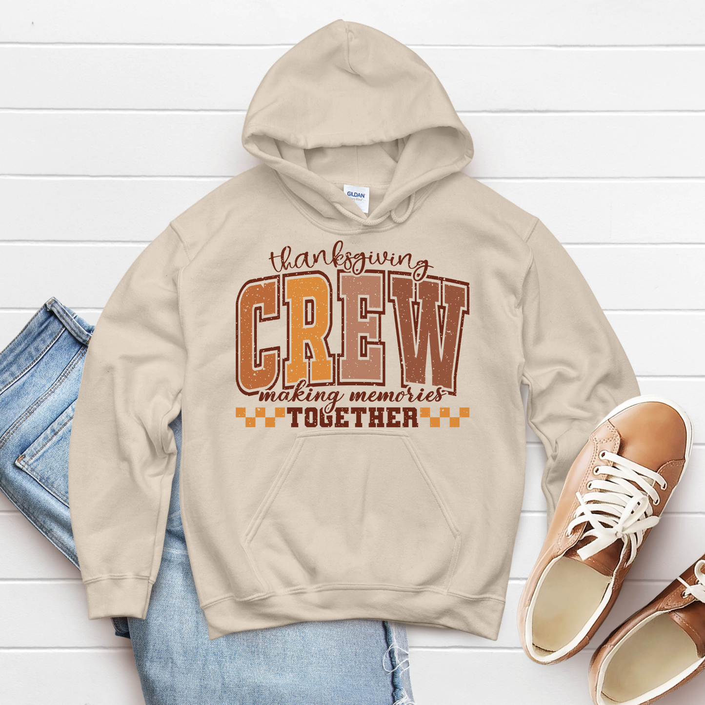 Beige hoodie with 'Thanksgiving Crew making memories together' text on a white wooden surface with jeans and shoes.