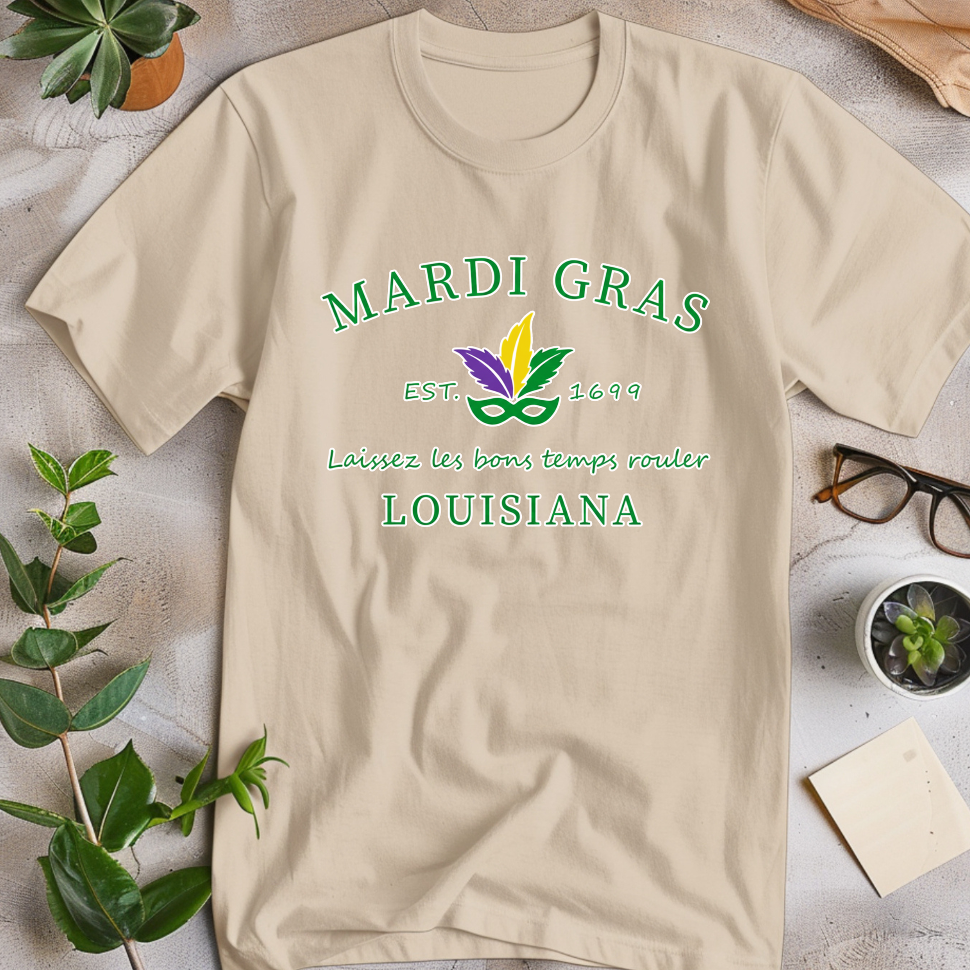 Beige t-shirt with Mardi Gras-themed design on a light surface with plants and glasses.