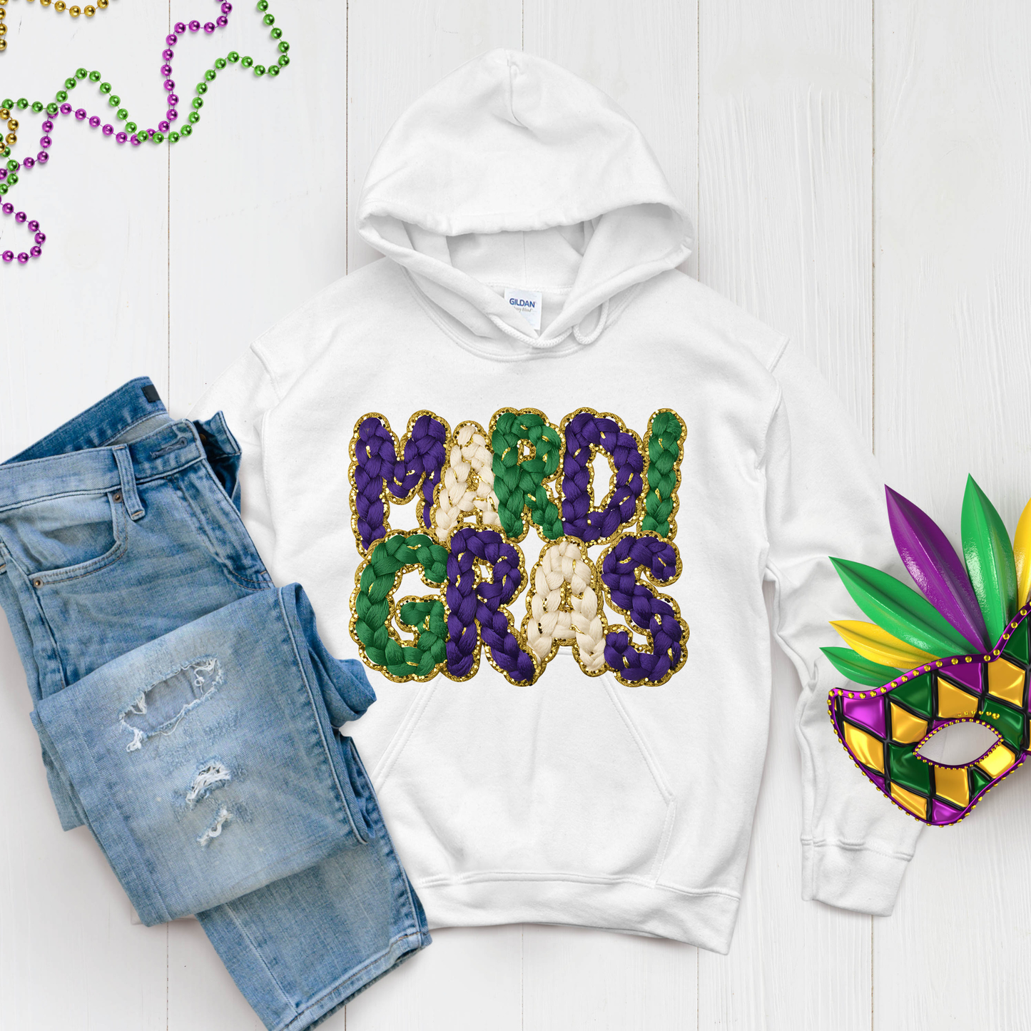 White hoodie with 'MARDI GRAS' text, blue jeans, and Mardi Gras mask on a light wooden surface.