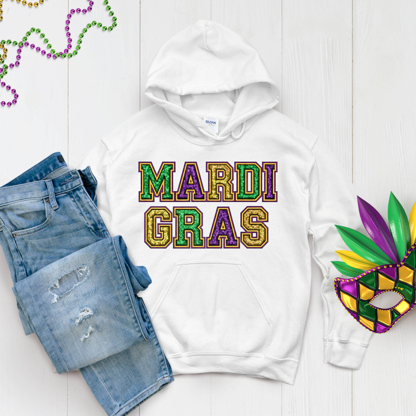 White hoodie with 'Mardi Gras' text, denim shorts, and Mardi Gras mask on a light wooden surface.