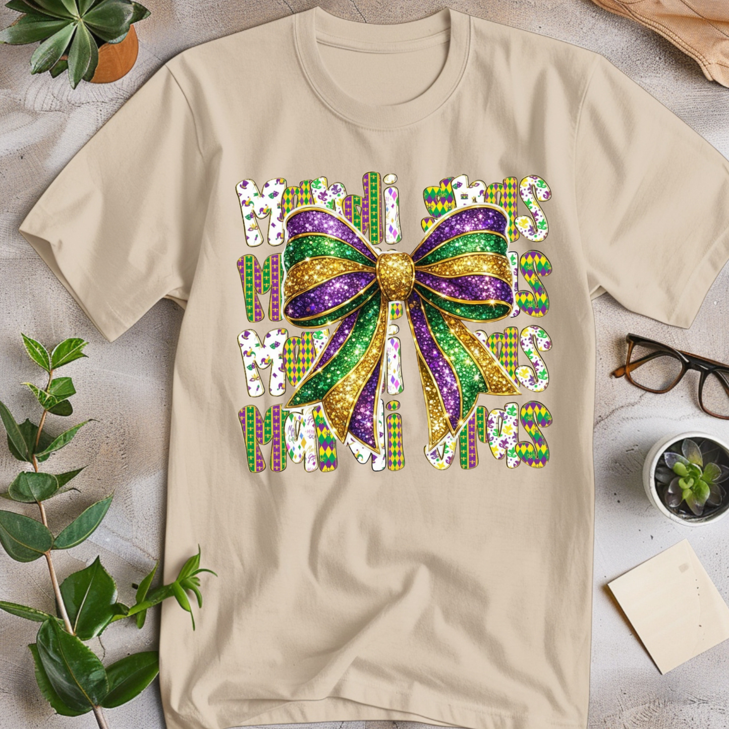 Beige t-shirt with colorful Mardi Gras-themed design on a light surface with plants and glasses.