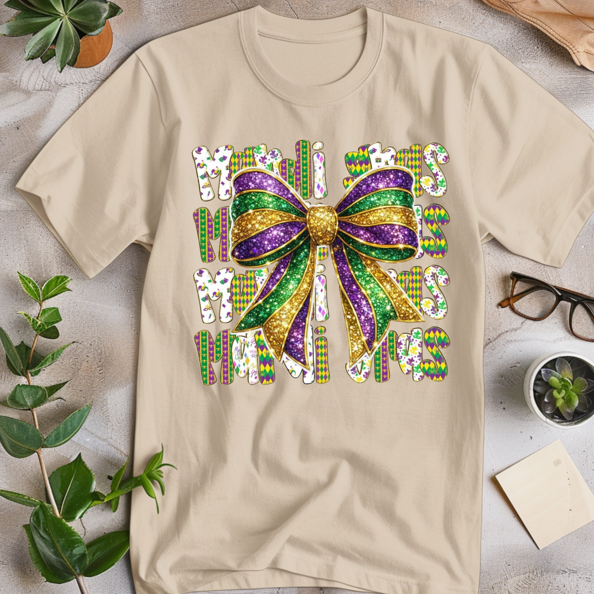 Beige t-shirt with colorful Mardi Gras-themed design on a light surface with plants and glasses.