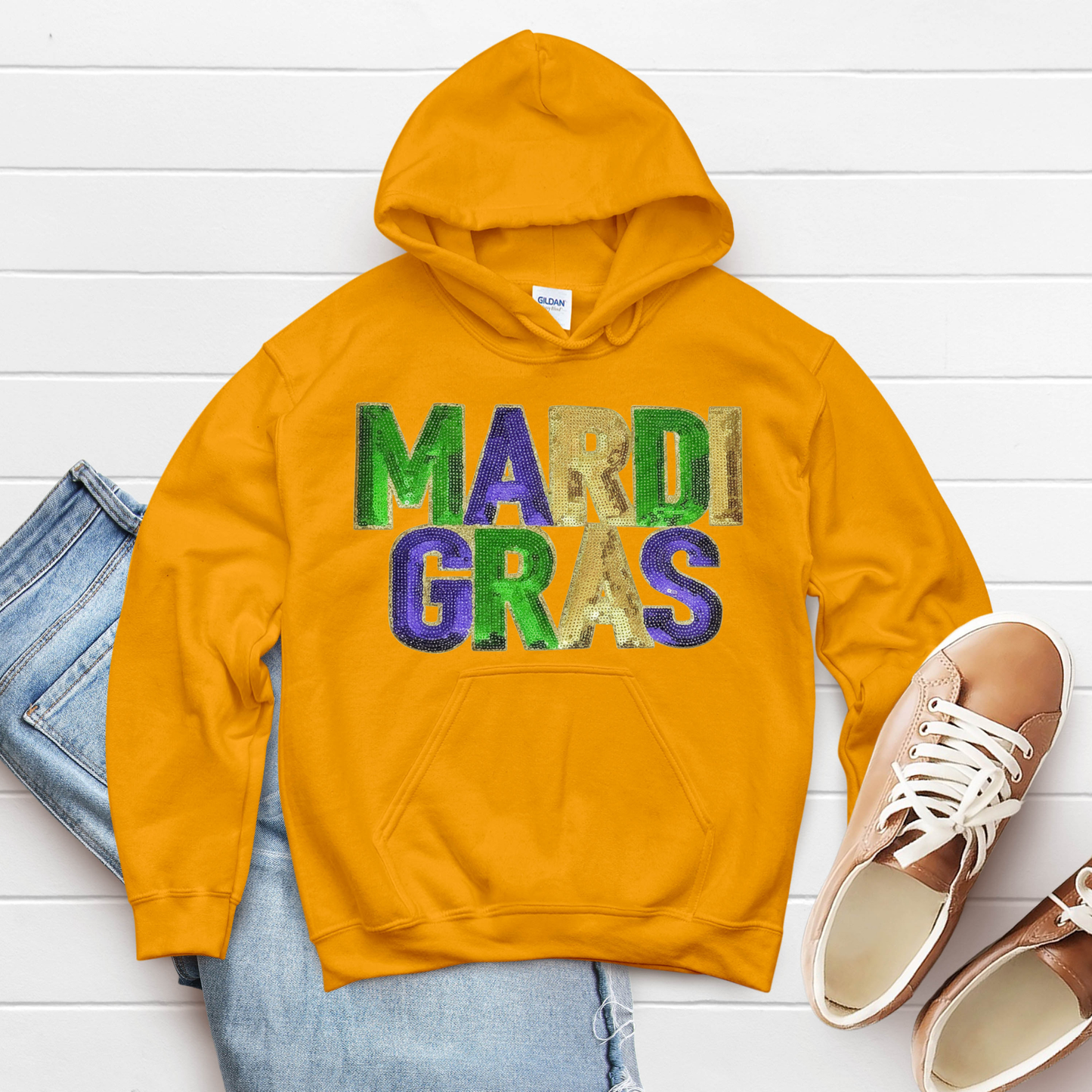 Yellow hoodie with 'MARDI GRAS' text, blue jeans, and brown shoes on a white wooden surface