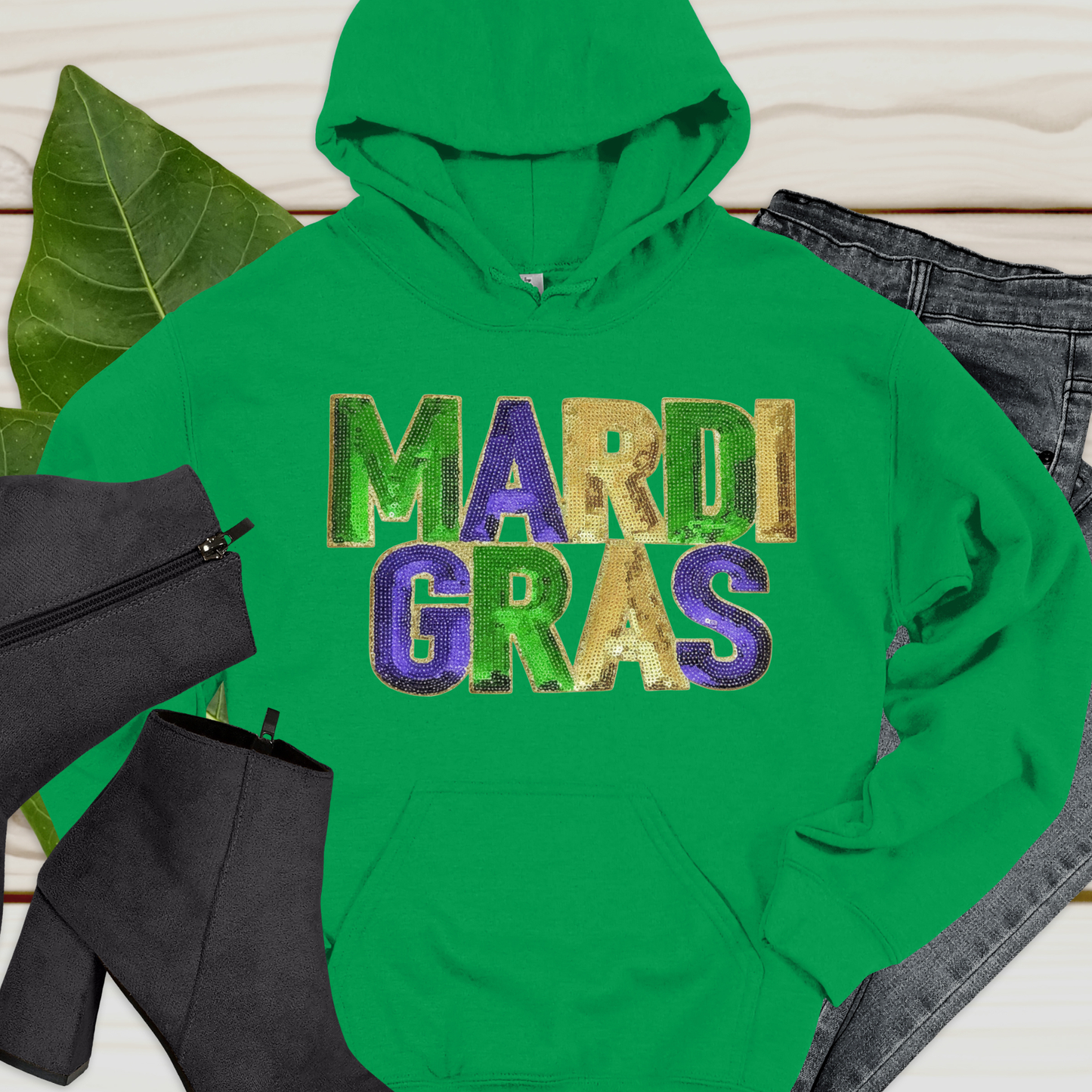 Green hoodie with 'Mardi Gras' text on a light wooden surface with leaves and black bag.