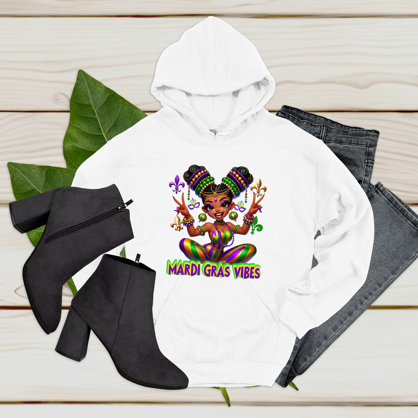 White hoodie with Mardi Gras-themed design, black boots, and gray jeans on a wooden surface.