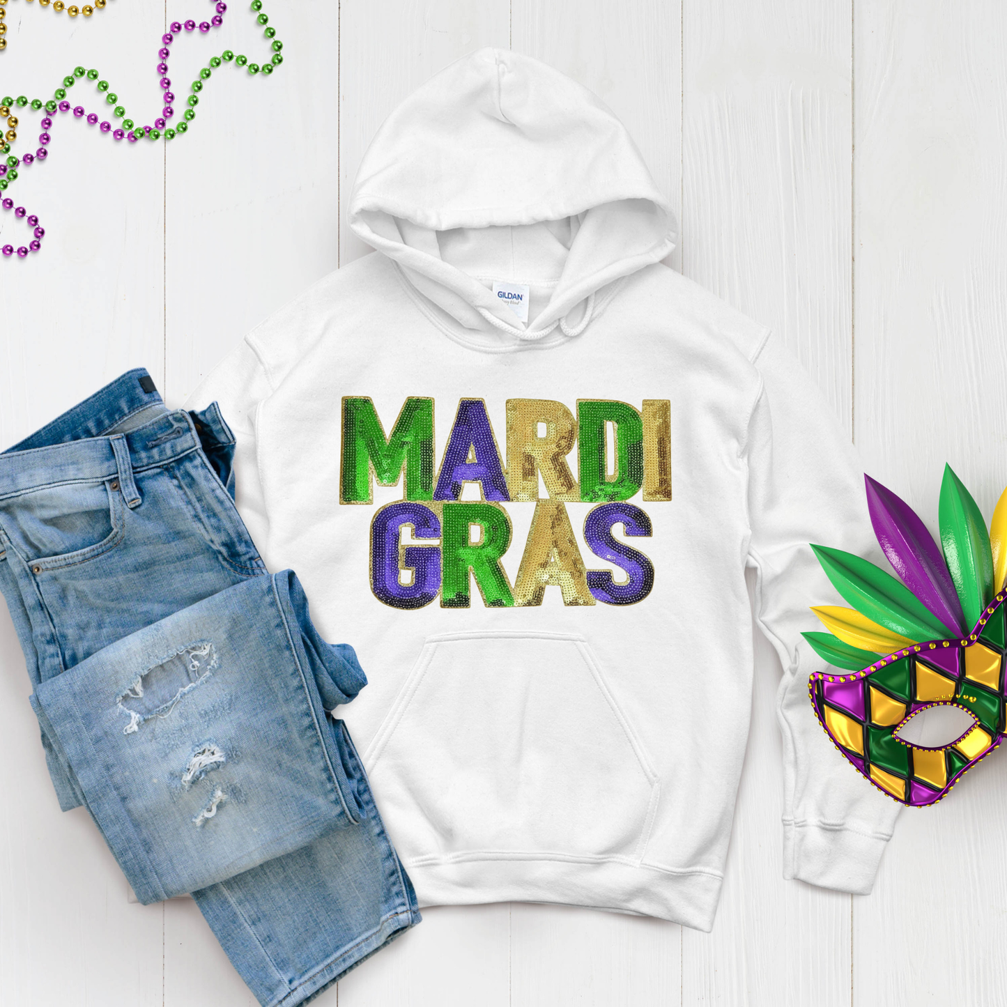 White hoodie with 'Mardi Gras' text, blue jeans, and a Mardi Gras mask on a light wooden surface.