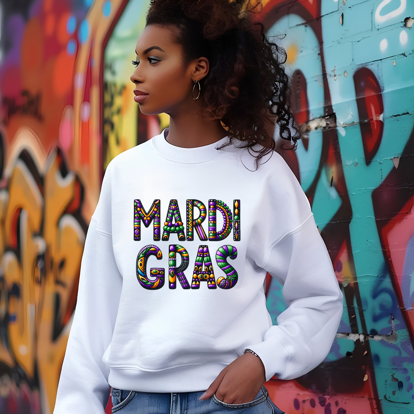 Person wearing a white sweatshirt with 'MARDI GRAS' in colorful letters against a graffiti-covered wall.