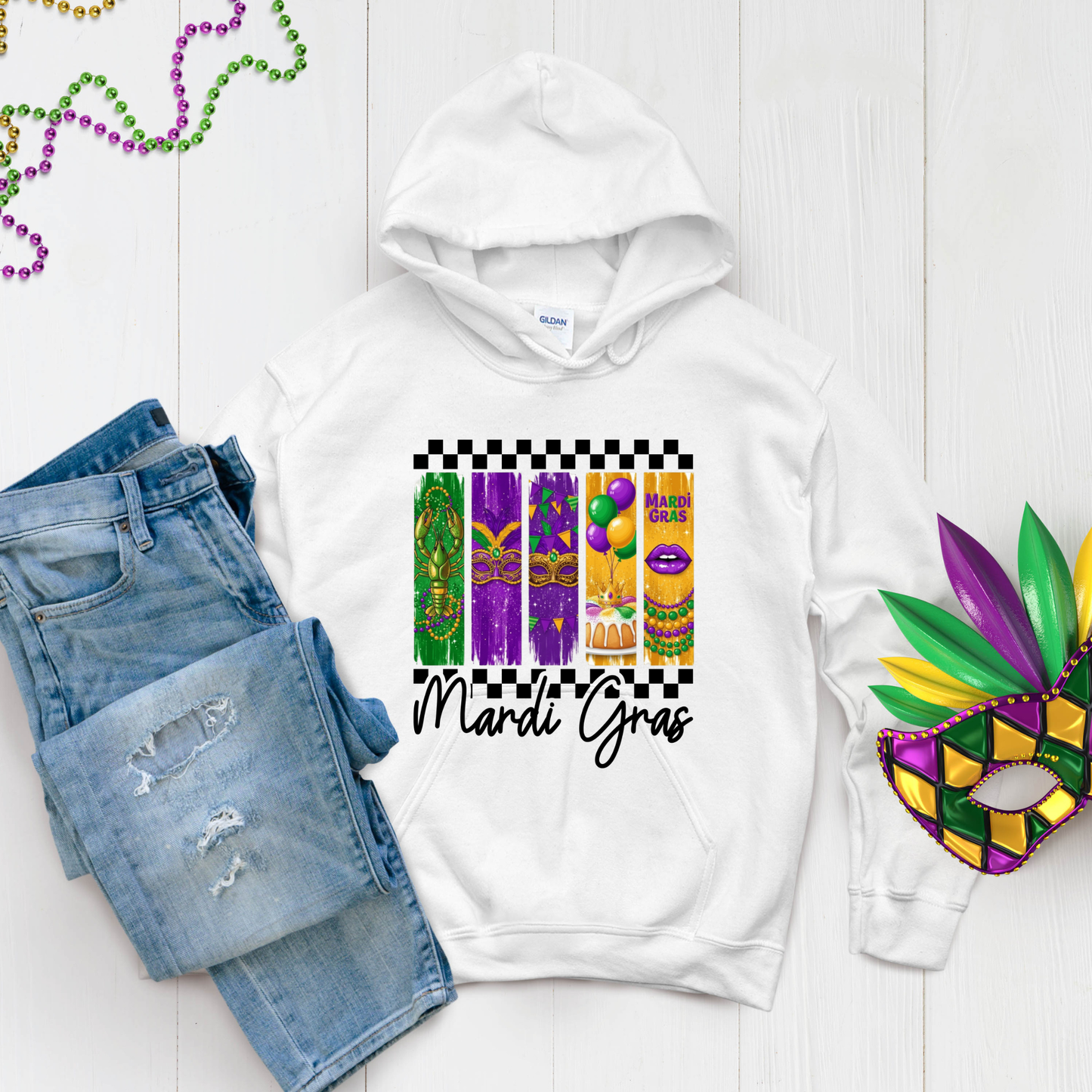 White hoodie with Mardi Gras-themed design, denim shorts, and a mask on a light wooden surface.