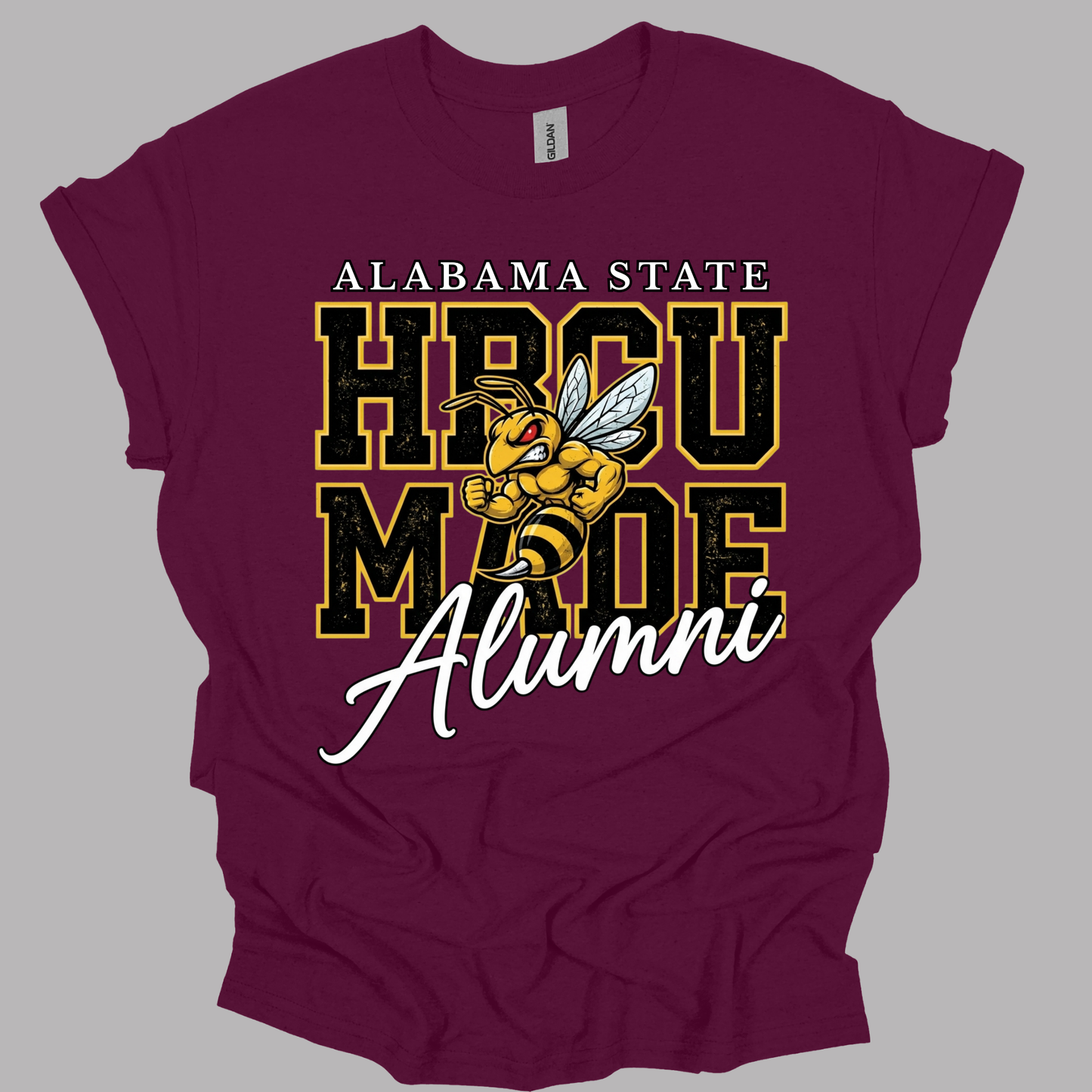 A maroon short-sleeve t-shirt with a graphic print reading 'ALABAMA STATE HBCU MADE Alumni' and featuring a yellow and black bee mascot, shown on a solid gray background.