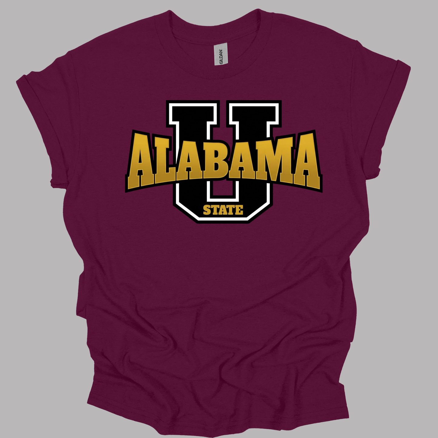 A maroon short-sleeve t-shirt with a graphic of a large black and white letter 'U' and the word 'ALABAMA' in gold block letters, with 'STATE' below, shown on a solid gray background.