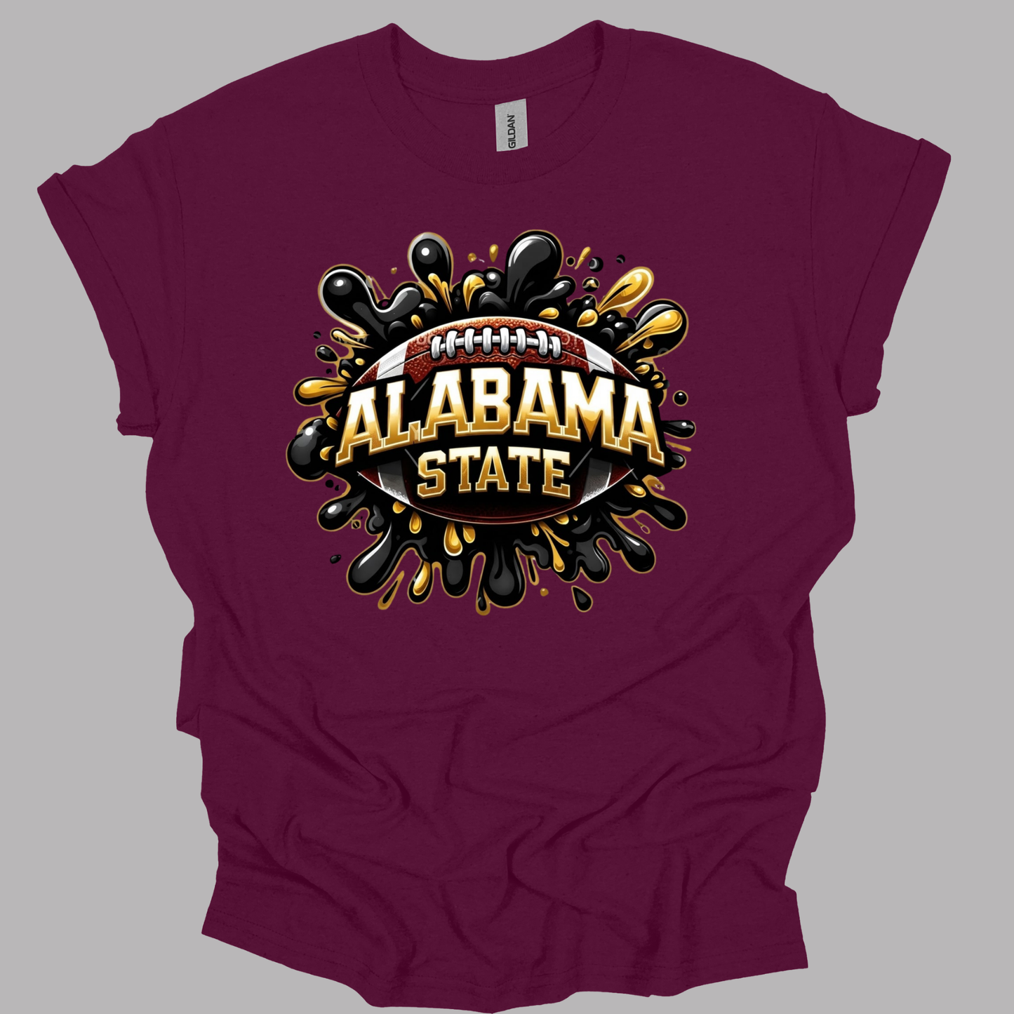 A maroon short-sleeve t-shirt with a graphic print on the front featuring the words 'ALABAMA STATE' in gold and white, set against a football and black/gold paint splatter design, displayed on a solid gray background.