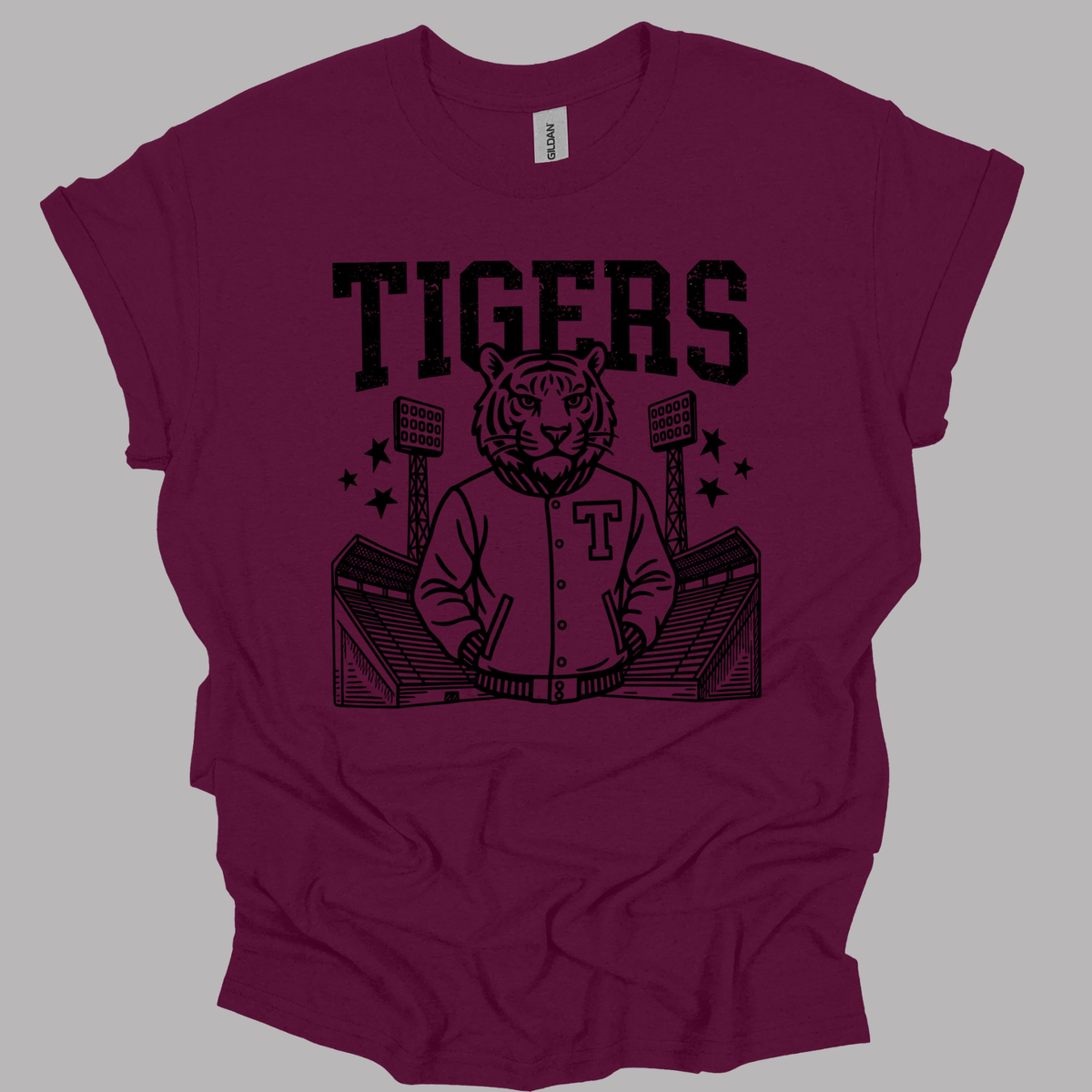 A maroon short-sleeve t-shirt with a black graphic print of a tiger mascot wearing a varsity jacket, seated in a stadium, with the word 'TIGERS' printed above. The shirt is displayed flat against a solid gray background.