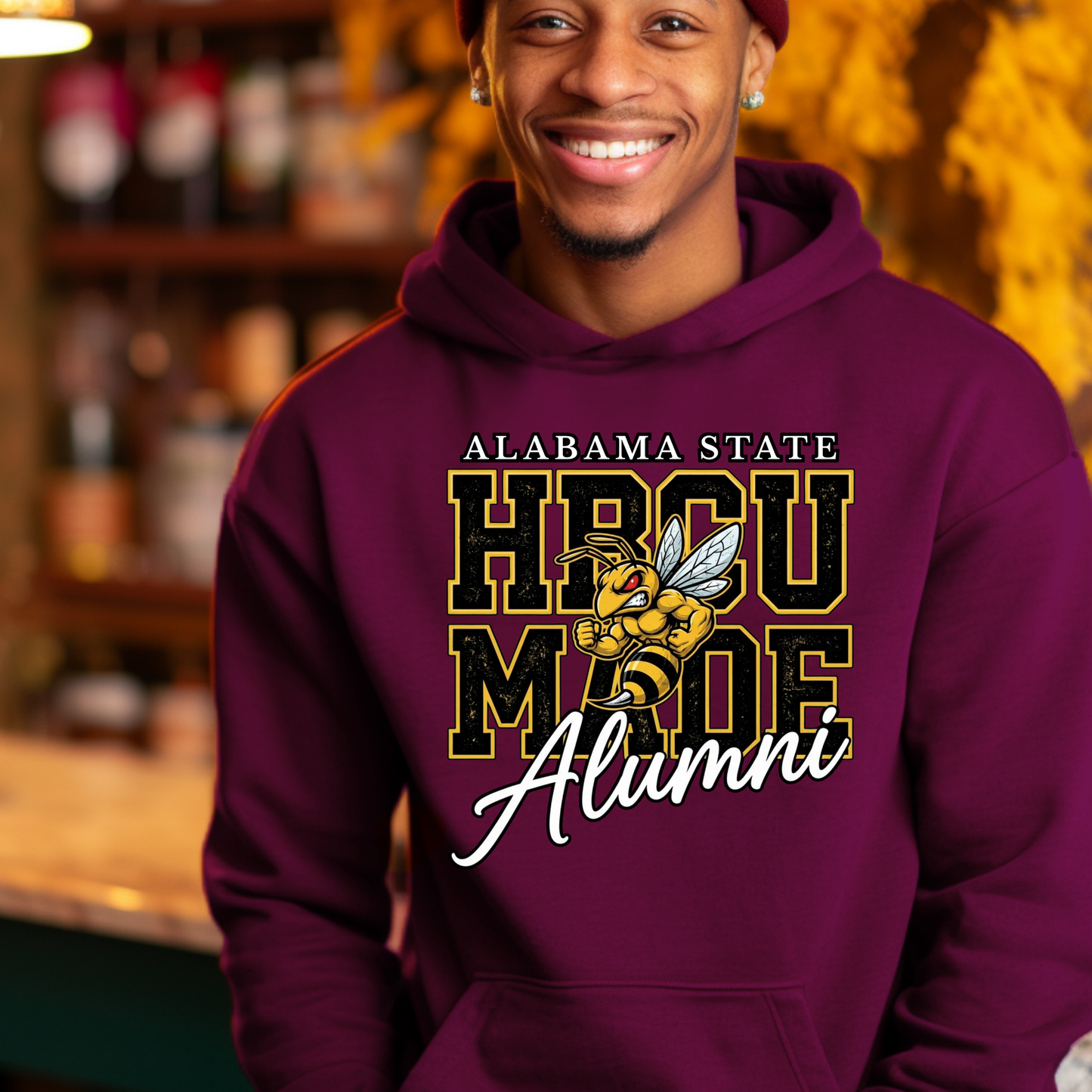 A smiling man wearing a maroon pullover hoodie with a graphic that reads 'ALABAMA STATE HBCU MADE ALUMNI' with a bee illustration, photographed in a warm, blurred indoor setting.