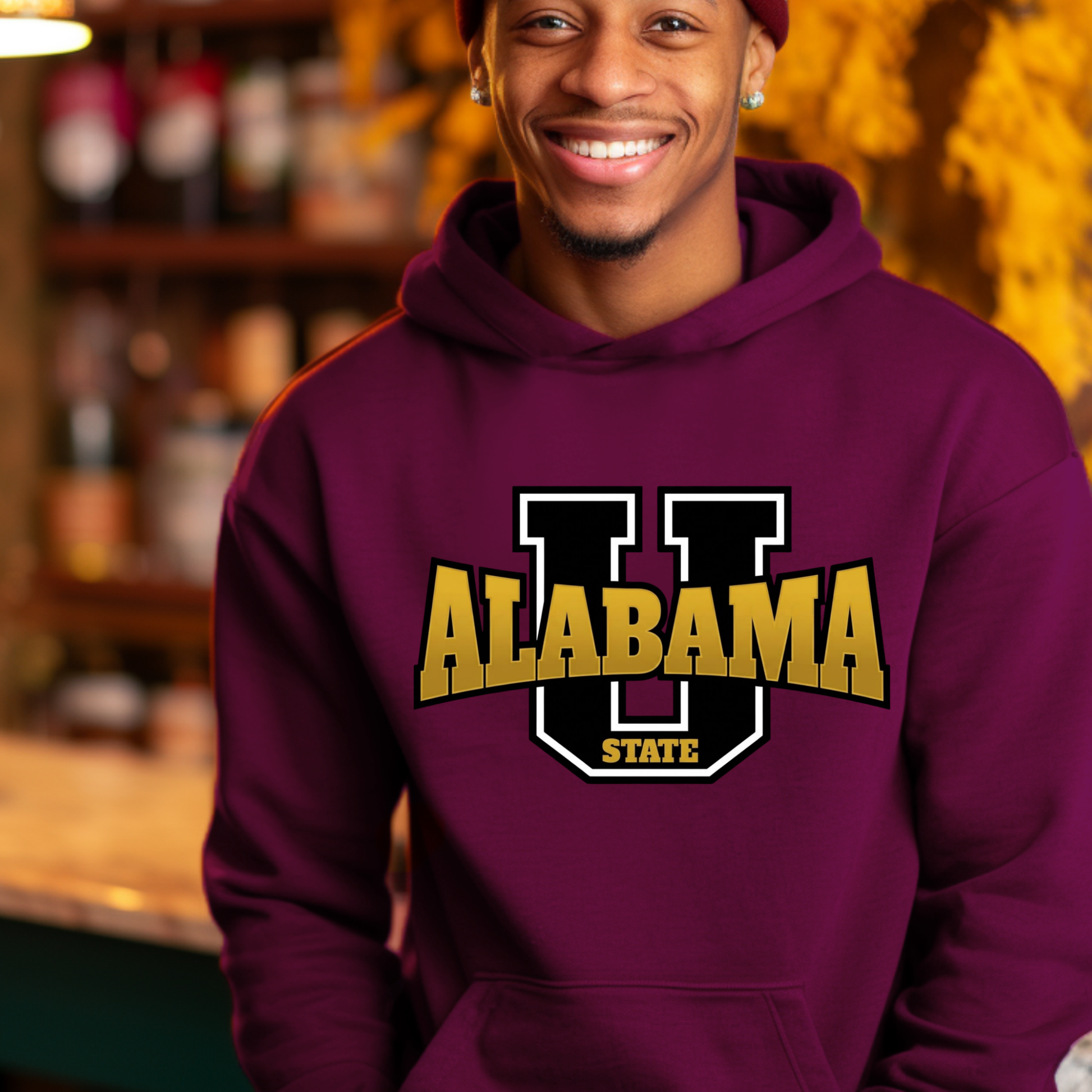 A smiling man wearing a maroon pullover hoodie with a graphic that reads 'ALABAMA STATE HBCU MADE ALUMNI' with a bee illustration, photographed in a warm, blurred indoor setting.