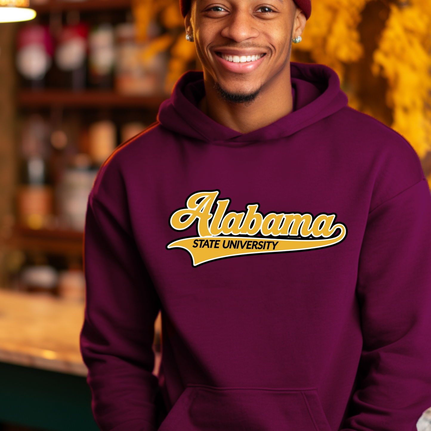 A smiling man wearing a maroon pullover hoodie with a graphic that reads 'ALABAMA STATE HBCU MADE ALUMNI' with a bee illustration, photographed in a warm, blurred indoor setting.