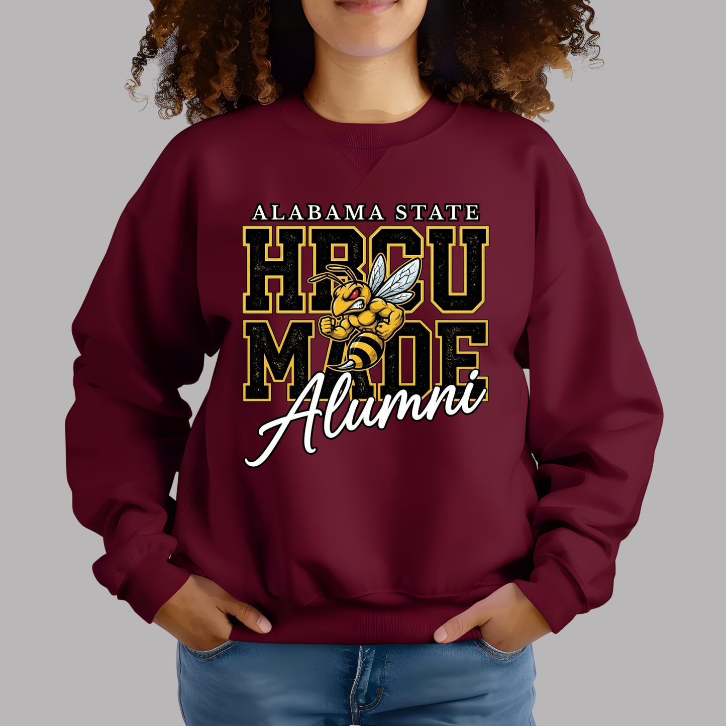 A woman wearing a maroon crewneck sweatshirt with a 'Alabama State HBCU Made Alumni' graphic on the front, paired with blue jeans, against a solid gray background.