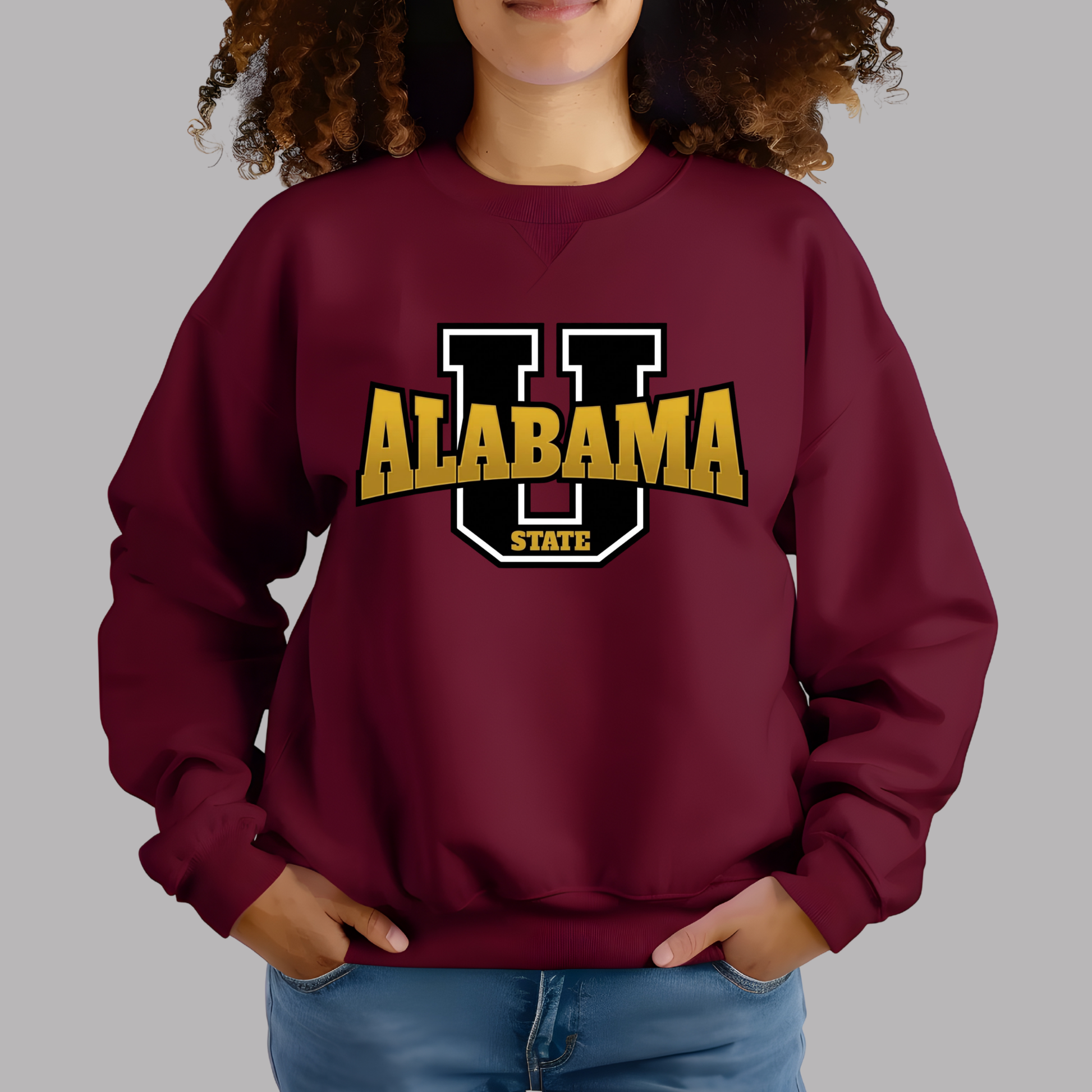 A woman wearing a maroon crewneck sweatshirt with a 'Alabama State HBCU Made Alumni' graphic on the front, paired with blue jeans, against a solid gray background.