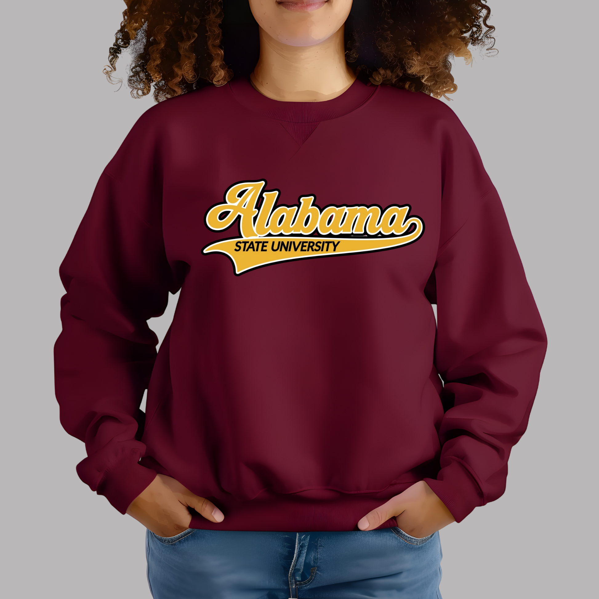 A woman wearing a maroon crewneck sweatshirt with a 'Alabama State HBCU Made Alumni' graphic in yellow, white, and black, paired with blue jeans against a solid gray background.