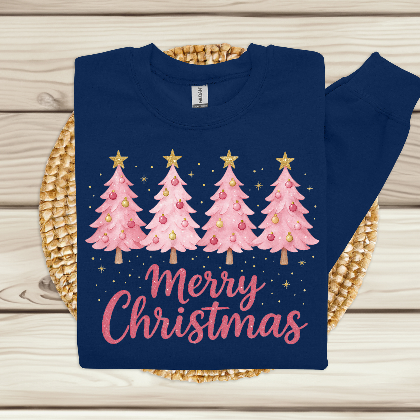 Folded navy blue sweatshirt with pink Christmas trees and 'Merry Christmas' text on a woven mat.