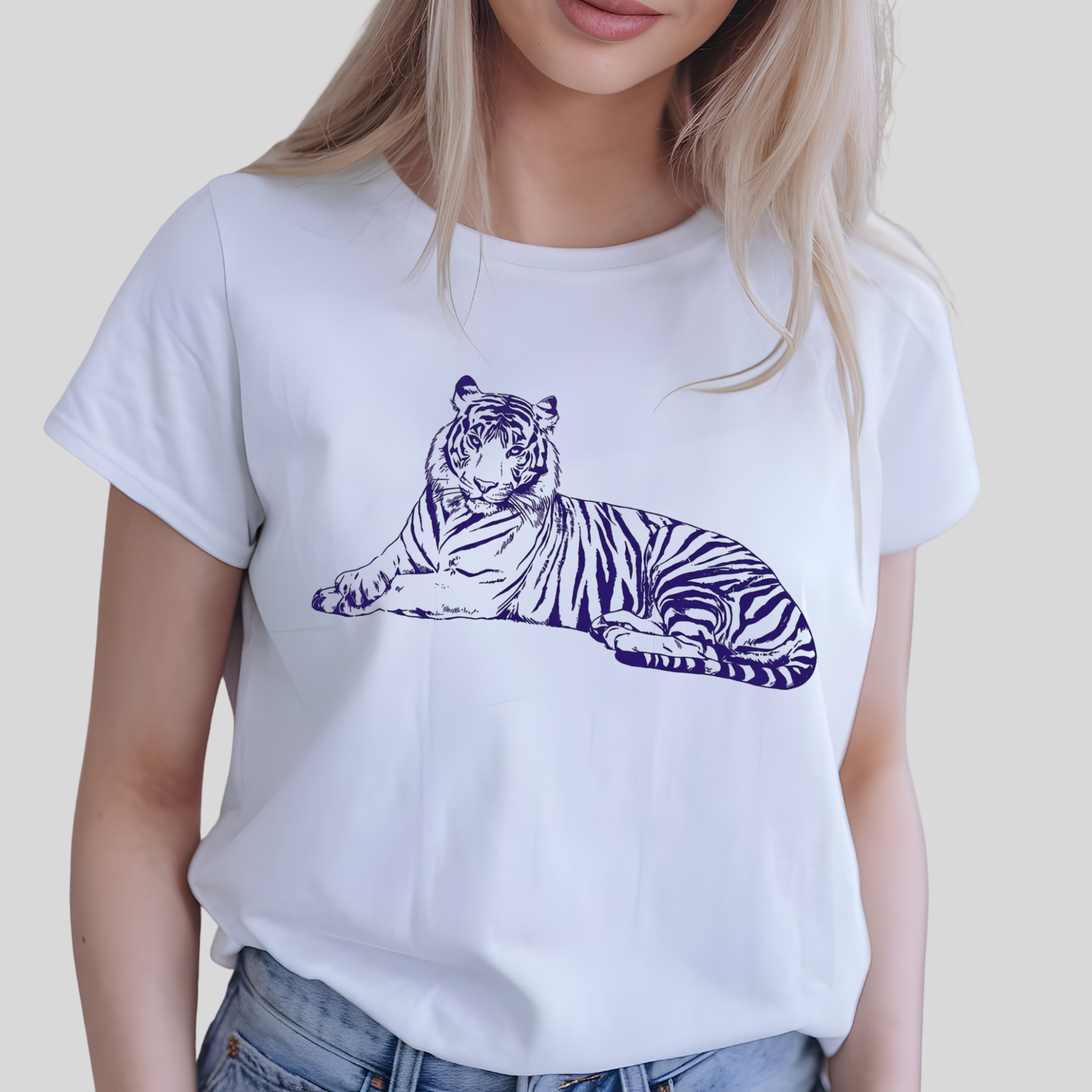 A woman wearing a white short-sleeve crewneck t-shirt with a dark blue line art graphic of a resting tiger on the front, paired with blue jeans against a light gray background.