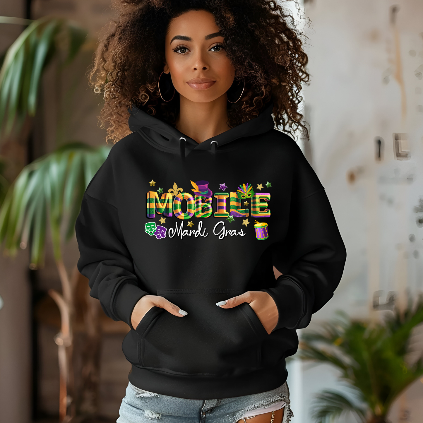 Person wearing a black hoodie with colorful 'Mardi Gras' text, standing indoors with plants in the background.