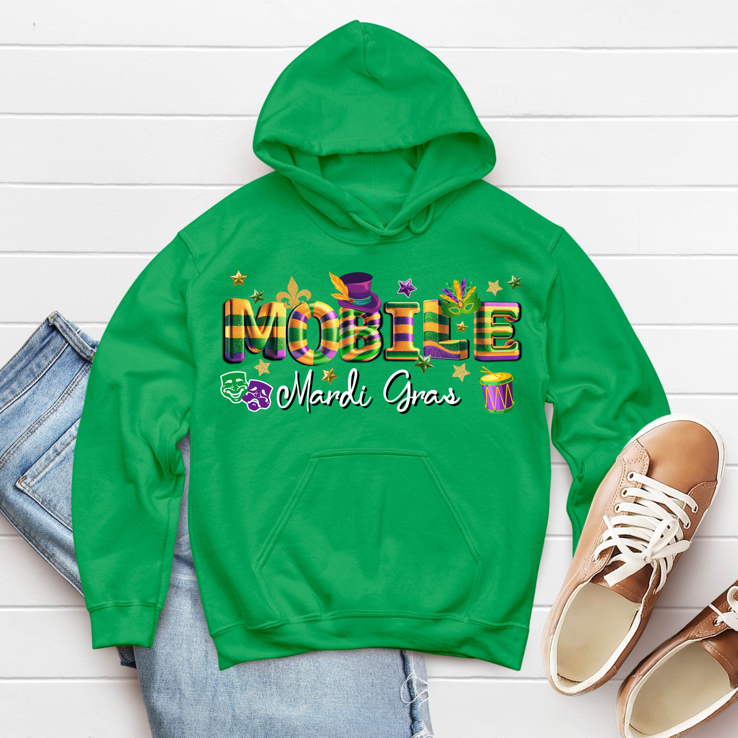 Green hoodie with 'Mardi Gras' text on a white wooden surface with jeans and shoes.