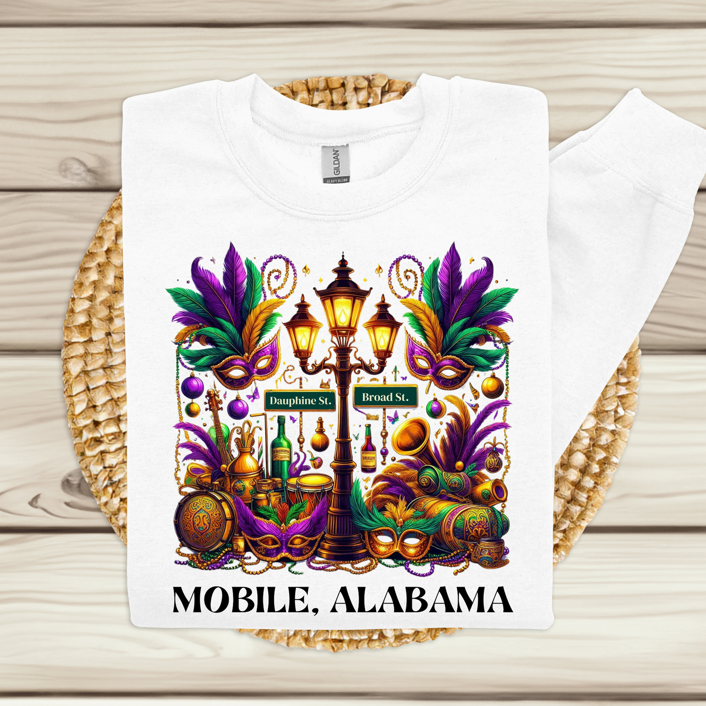 White t-shirt with Mardi Gras-themed design and 'Mobile, Alabama' text on a wooden surface.