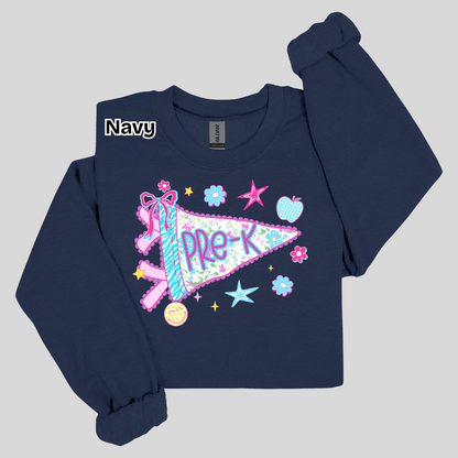 A navy blue long-sleeve sweatshirt with a colorful 'PRE-K' pennant graphic on the front, featuring a ribbon, stars, and flowers. The brand 'Gildan' is visible on the inner neck label, and a 'Navy' color label is overlaid on the upper left chest.