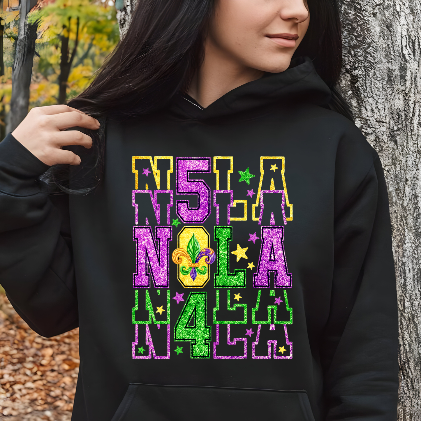 Person wearing a black hoodie with colorful 'NOLA' text and Mardi Gras elements, standing outdoors.