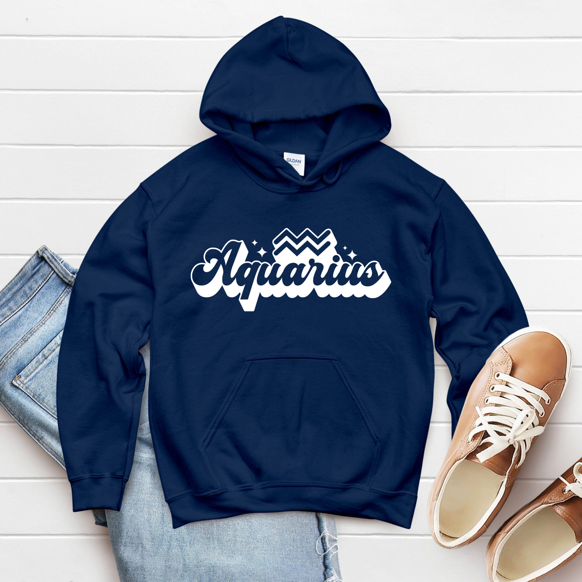 A navy blue pullover hoodie with 'Aquarius' in white script across the chest, styled in a flat lay with light blue jeans and tan sneakers on a white wood plank background.