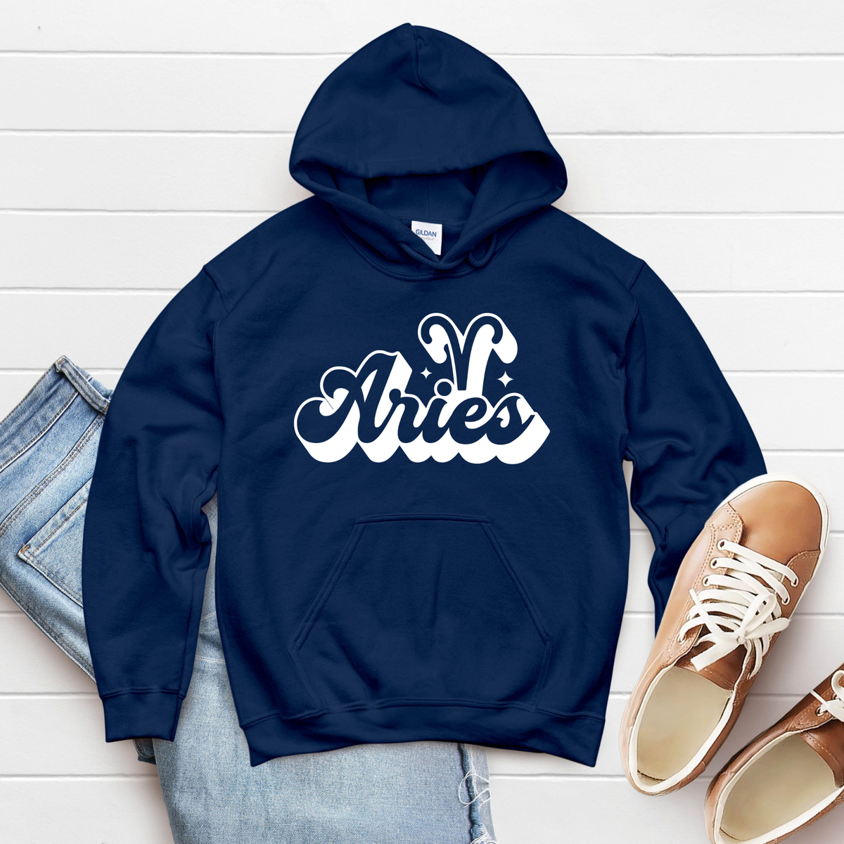 A navy blue hoodie with a white 'Aries' zodiac graphic, styled in a flat lay with light blue jeans and tan sneakers on a white wood plank background.