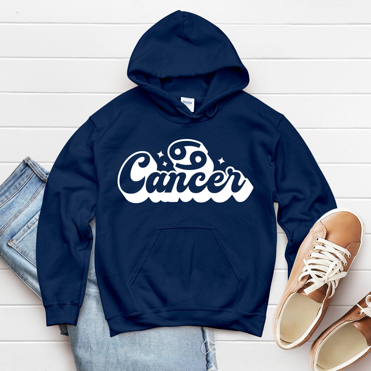 A navy blue hoodie with a white 'Cancer' zodiac graphic, styled in a flat lay with light blue jeans and tan sneakers on a white wood plank background.