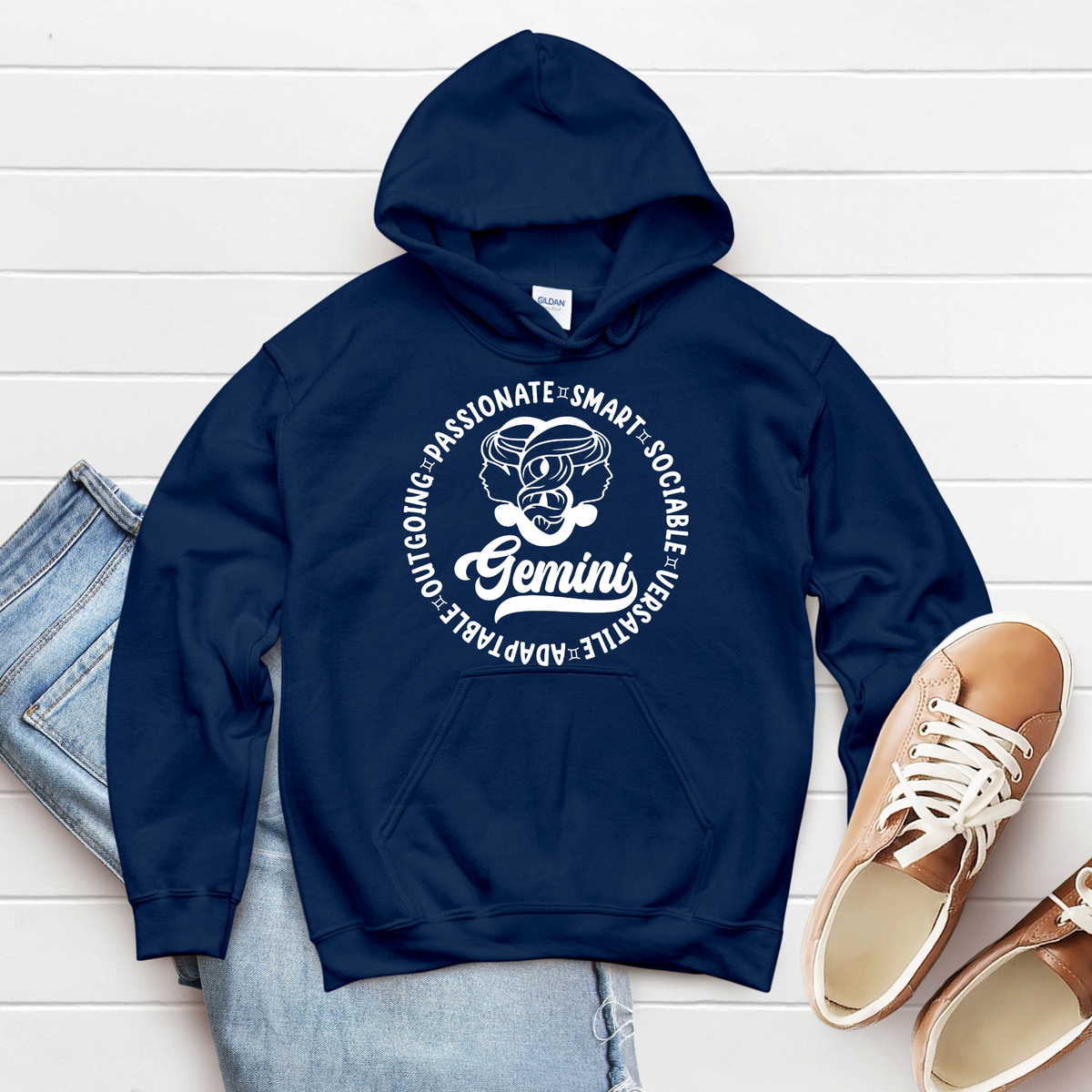 A navy blue hoodie with a white circular Gemini graphic print, styled in a flat lay with light-wash jeans and tan sneakers on a white wood plank background.