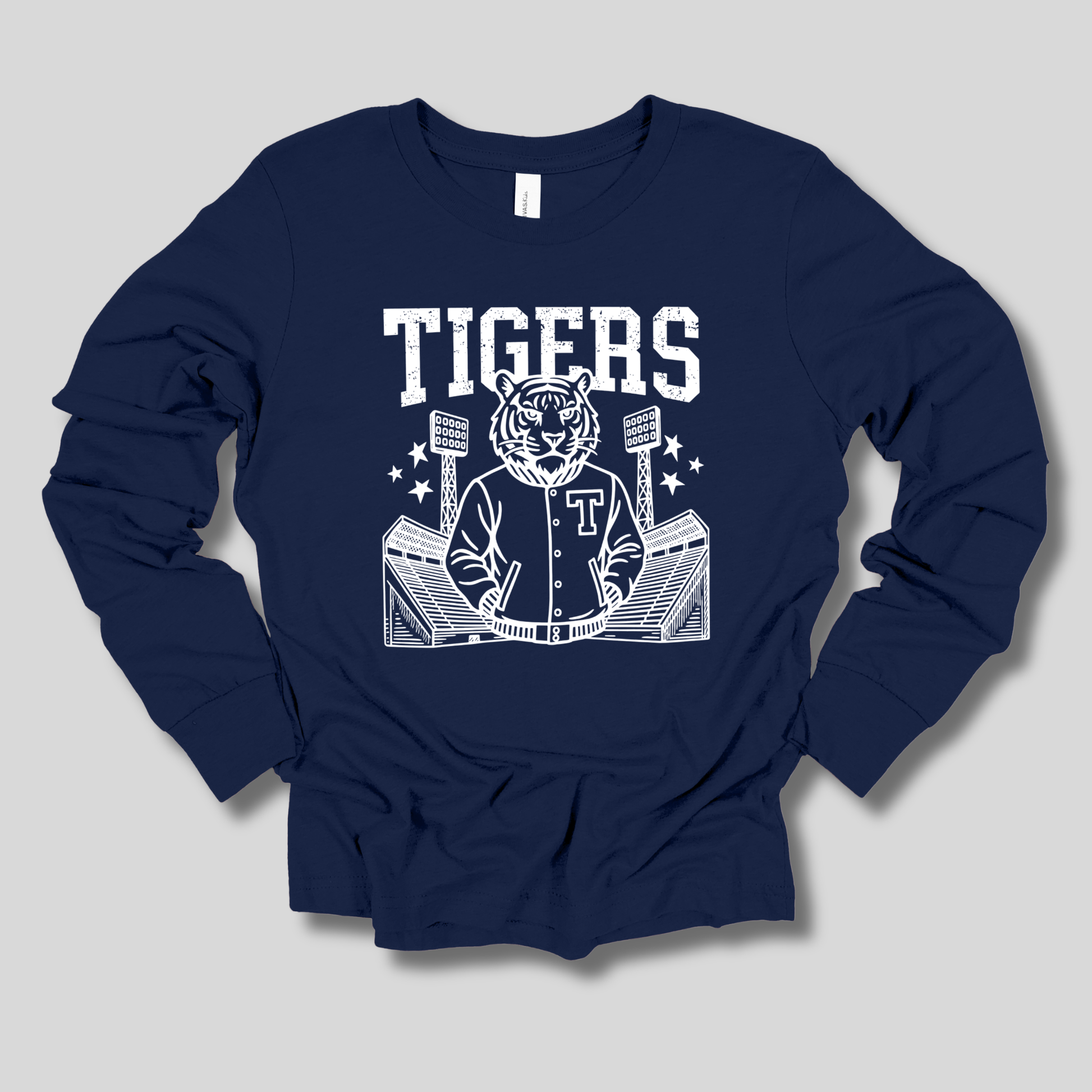 A navy blue long-sleeve t-shirt with a white graphic print on the front featuring the word 'TIGERS', a tiger mascot in a varsity jacket, and a stadium illustration, displayed on a neutral gray background.
