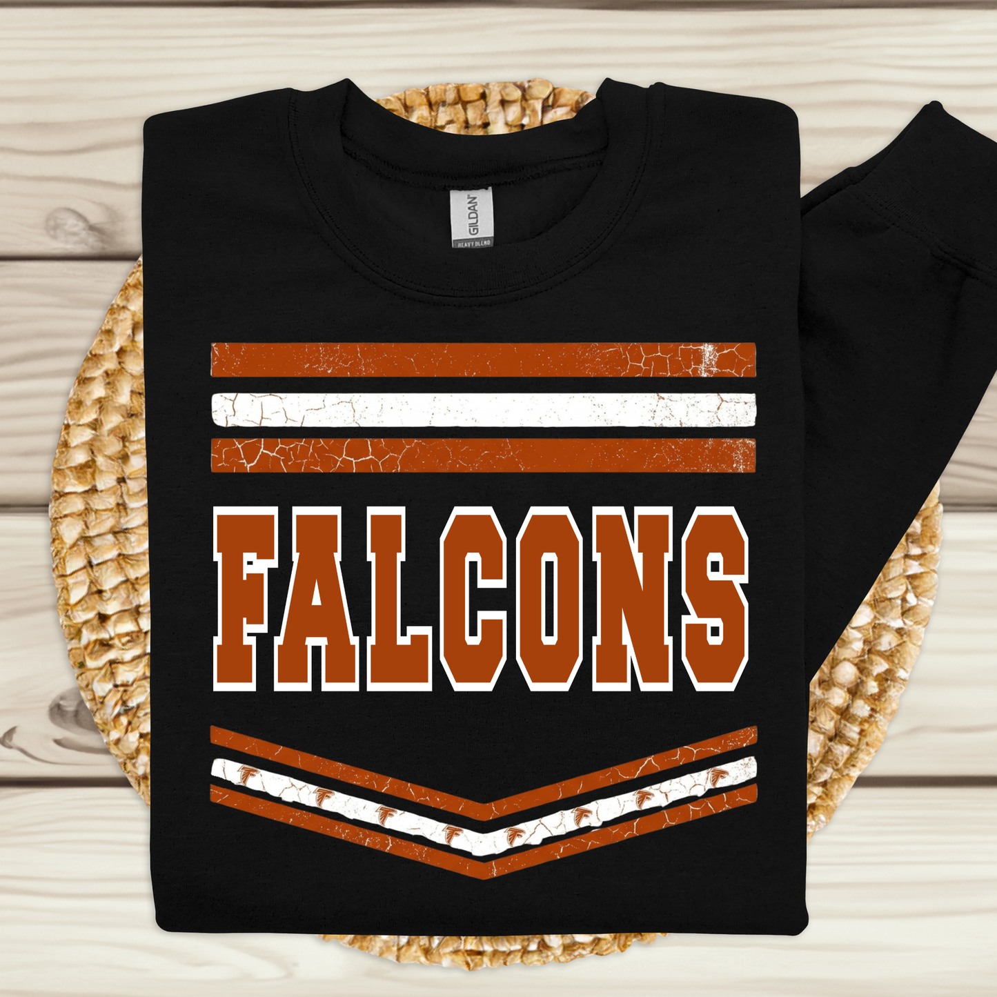 A black long-sleeve sweatshirt laid flat on a woven round mat over a light wood surface. The sweatshirt has a distressed orange and white graphic with the word 'FALCONS' across the front.
